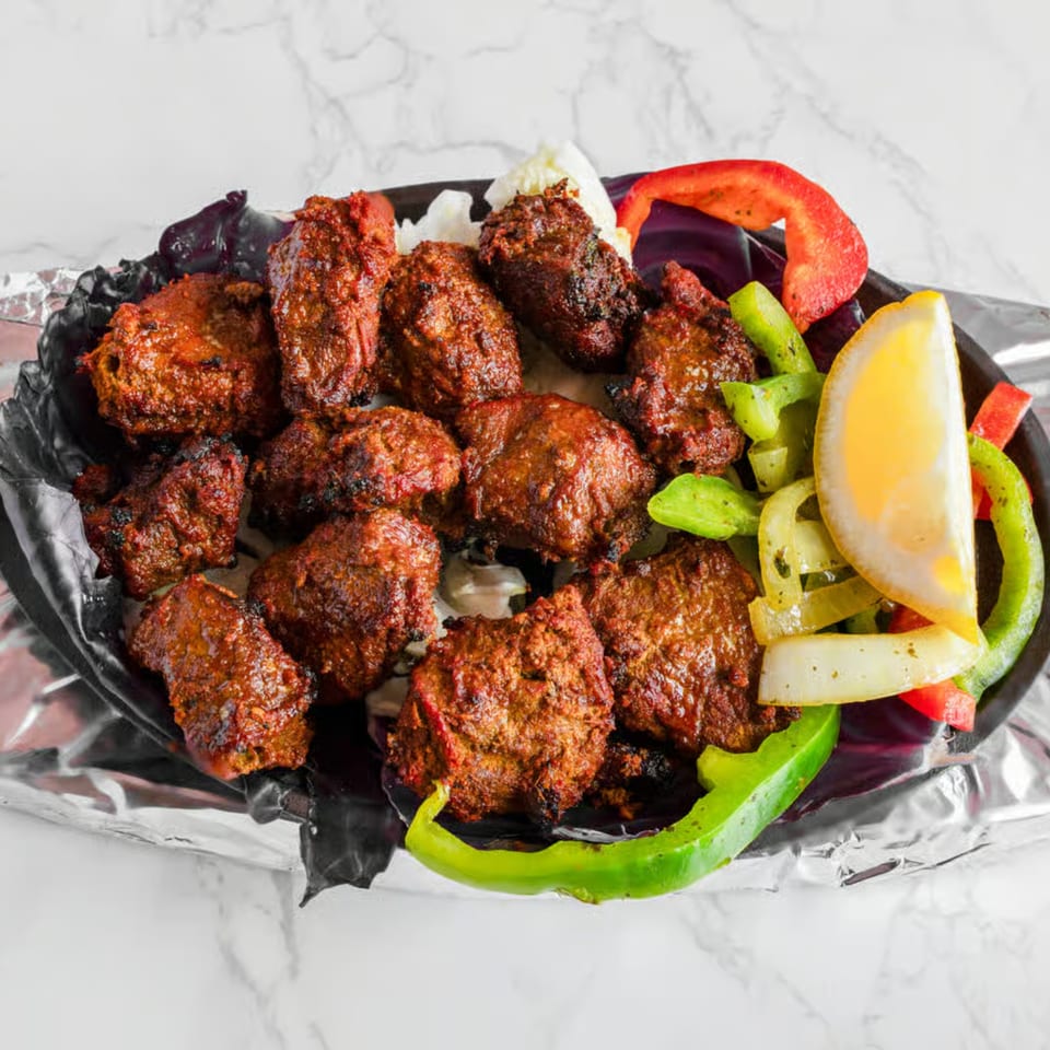 Indian lamb tikka, garnished with bell peppers, onions, and a lemon wedge, served on foil.