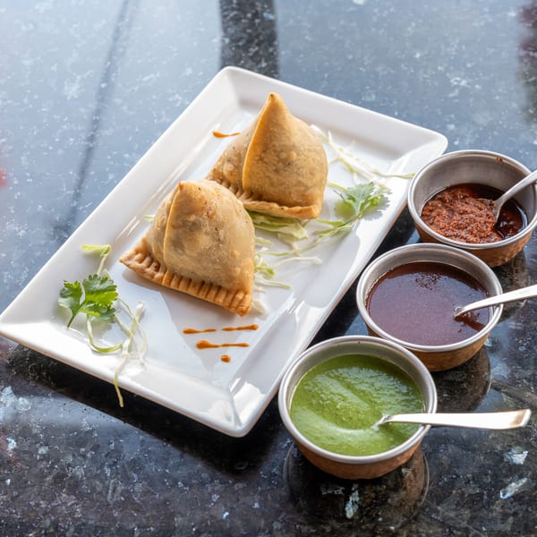 Two golden-brown samosas on a white plate with cilantro, served with three dipping sauces.