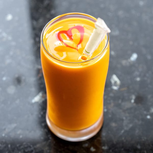 A vibrant orange mango lassi in a glass, topped with a red heart design and a straw.